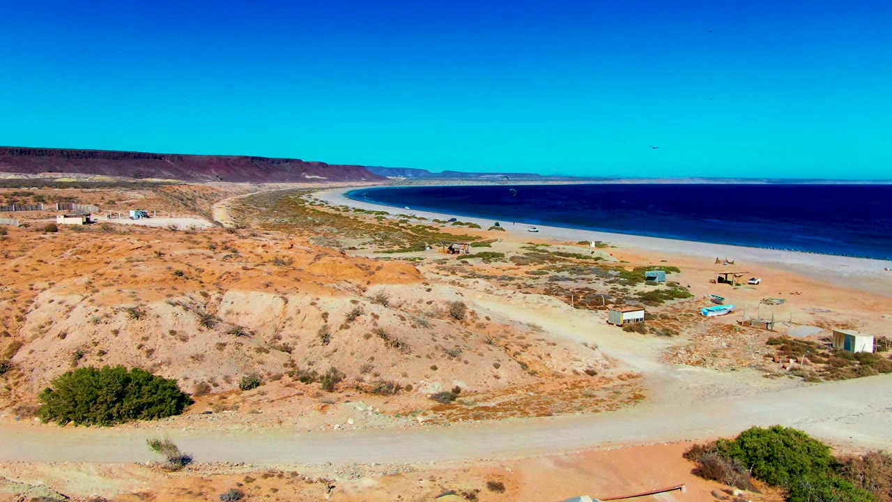 Wide aerial view over San Juanico, Baja California Sur, with a pale dirt road in the foreground, dry scrubland, sandy beach, dark blue ocean, distant hills, and no visible people.