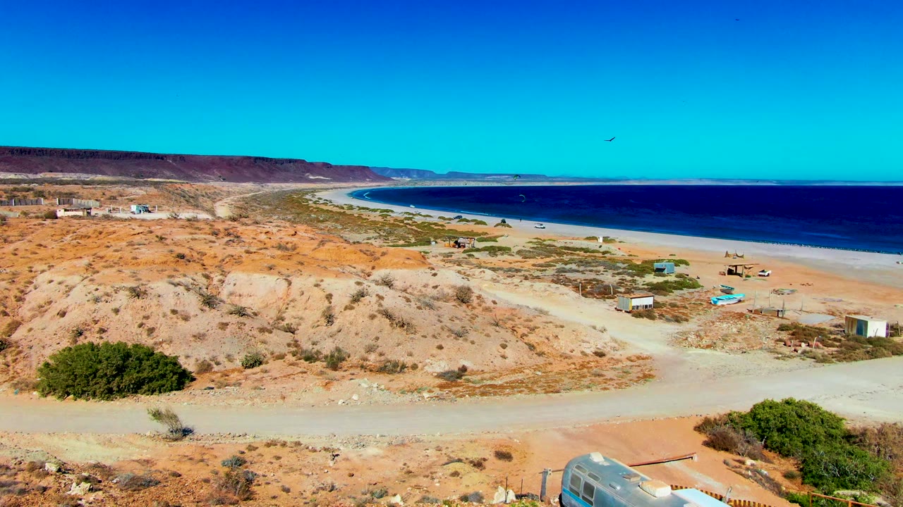 San Juanico aerial view with tan scrubland, a pale road crossing the lower frame, small structures near the beach, a long blue ocean horizon, and no visible people.