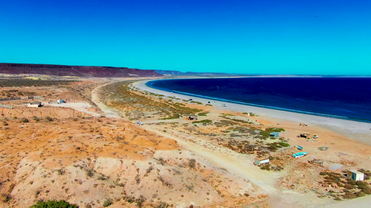 Aerial frame from San Juanico, Baja California Sur showing a pale dirt road across dry scrubland above a sandy beach and dark blue ocean, with no visible people.