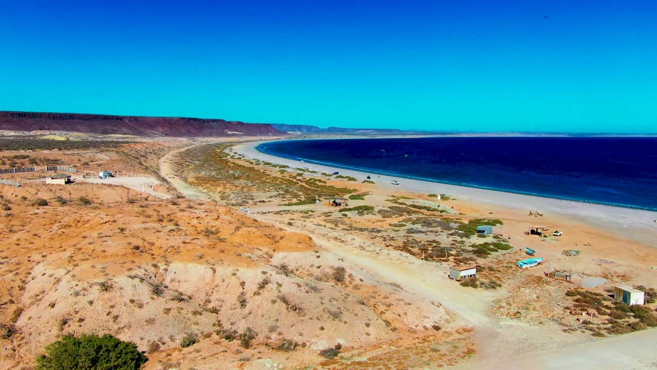 Drone-height view in San Juanico showing the dirt road and scrubby foreground leading toward beach shacks, pale sand, and a dark blue ocean band, with no visible people.