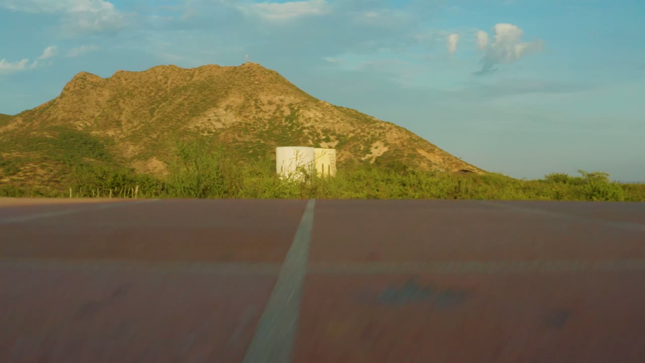 At 0:00, a reddish road fills the lower foreground in Cabo Pulmo, with green roadside vegetation, white cylindrical tanks, a rocky mountain ridge, scattered clouds, and no visible people.