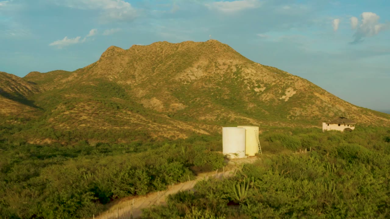 At 0:02, the frame widens across the Cabo Pulmo hillside, placing the white tanks near the lower center, a dirt track through scrub, a pale structure on the right slope, and no visible people.