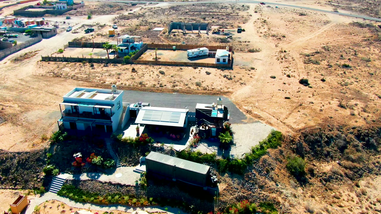 Aerial frame over a San Juanico desert property with modern buildings, a driveway, fenced lots, a white trailer in the background, sparse brush, and no visible people.