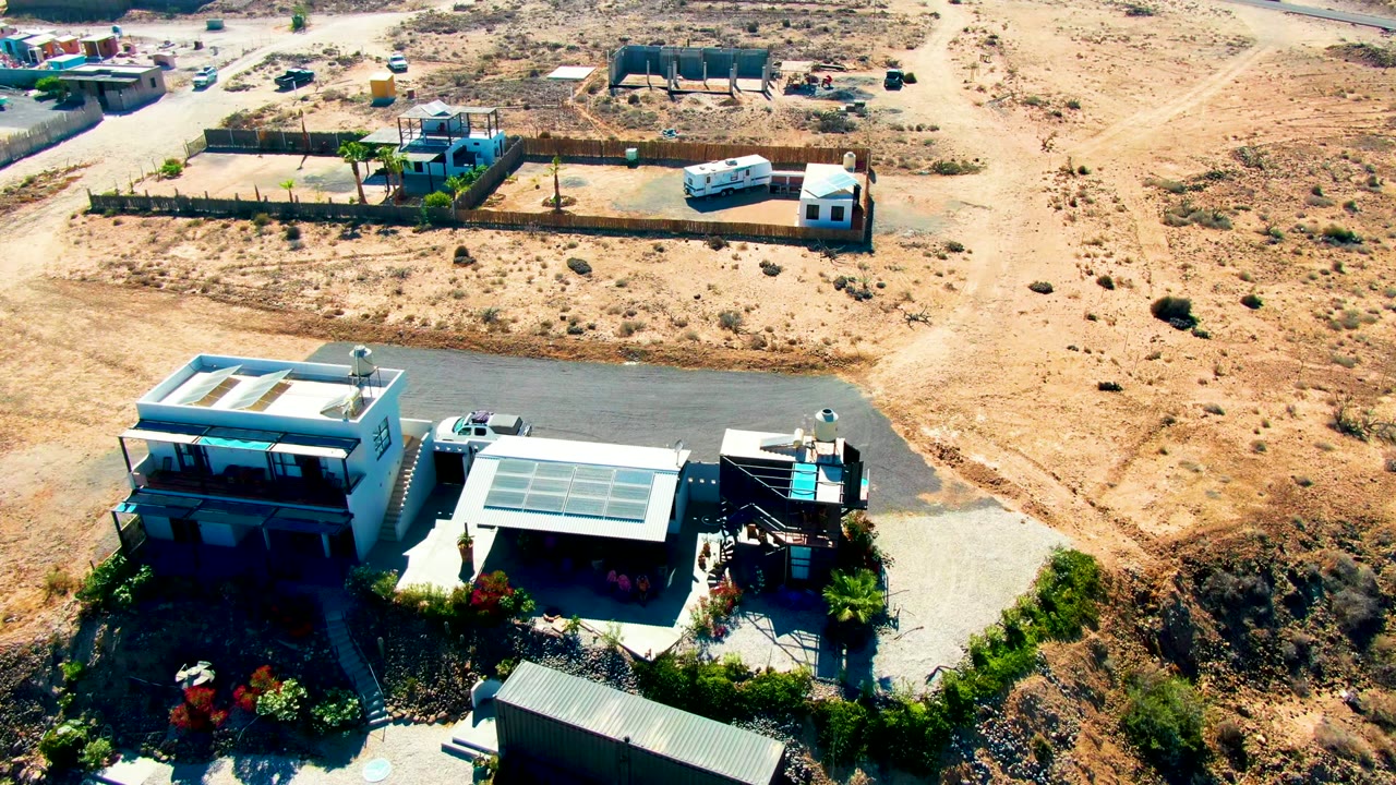 Drone view over a fenced desert property with buildings, road, and trailer.