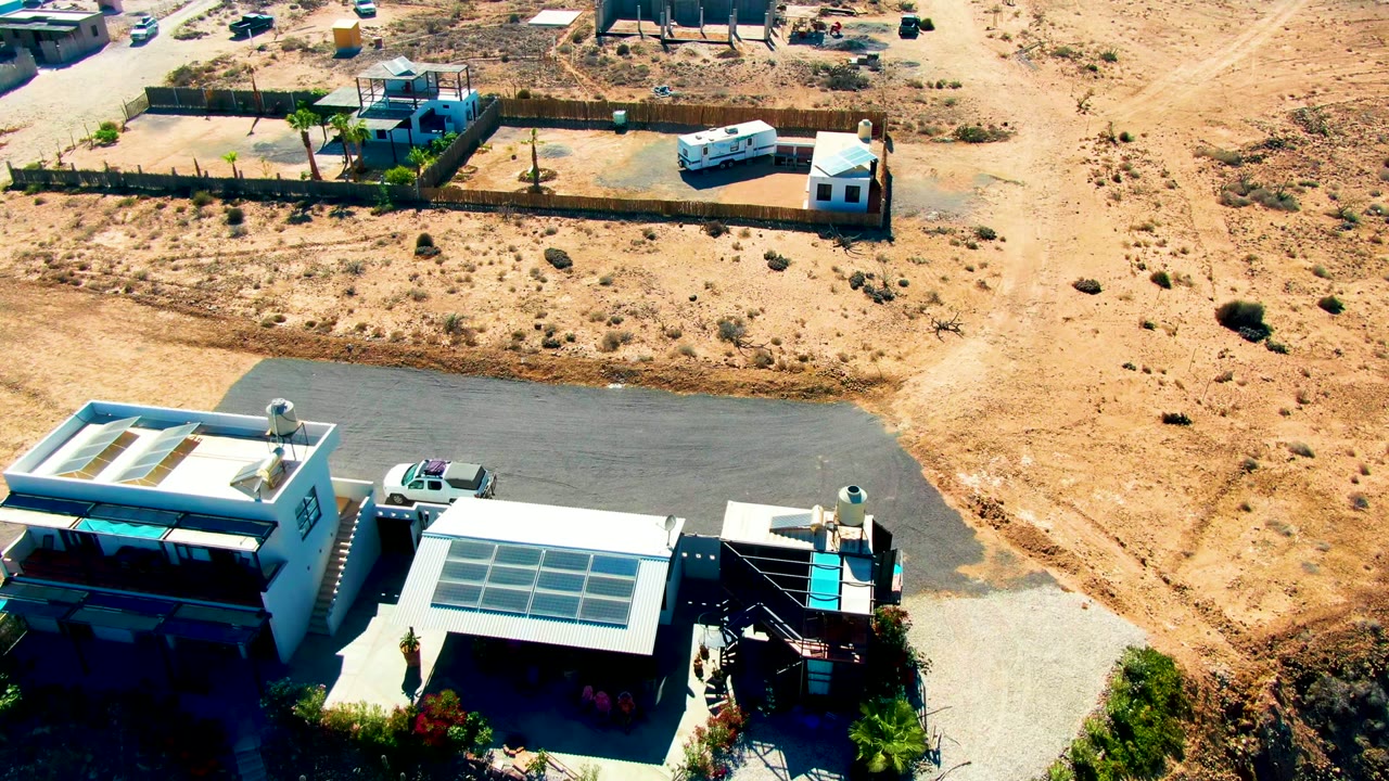 High-angle San Juanico frame with shaded building roofs at lower right, a paved or packed driveway, fenced sandy lots, scattered desert vegetation, and no visible people.