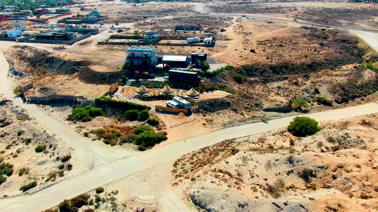 Wide aerial frame over San Juanico, Baja California Sur showing a small desert camp, low buildings, tents, sandy roads, sparse shrubs, and no visible people.