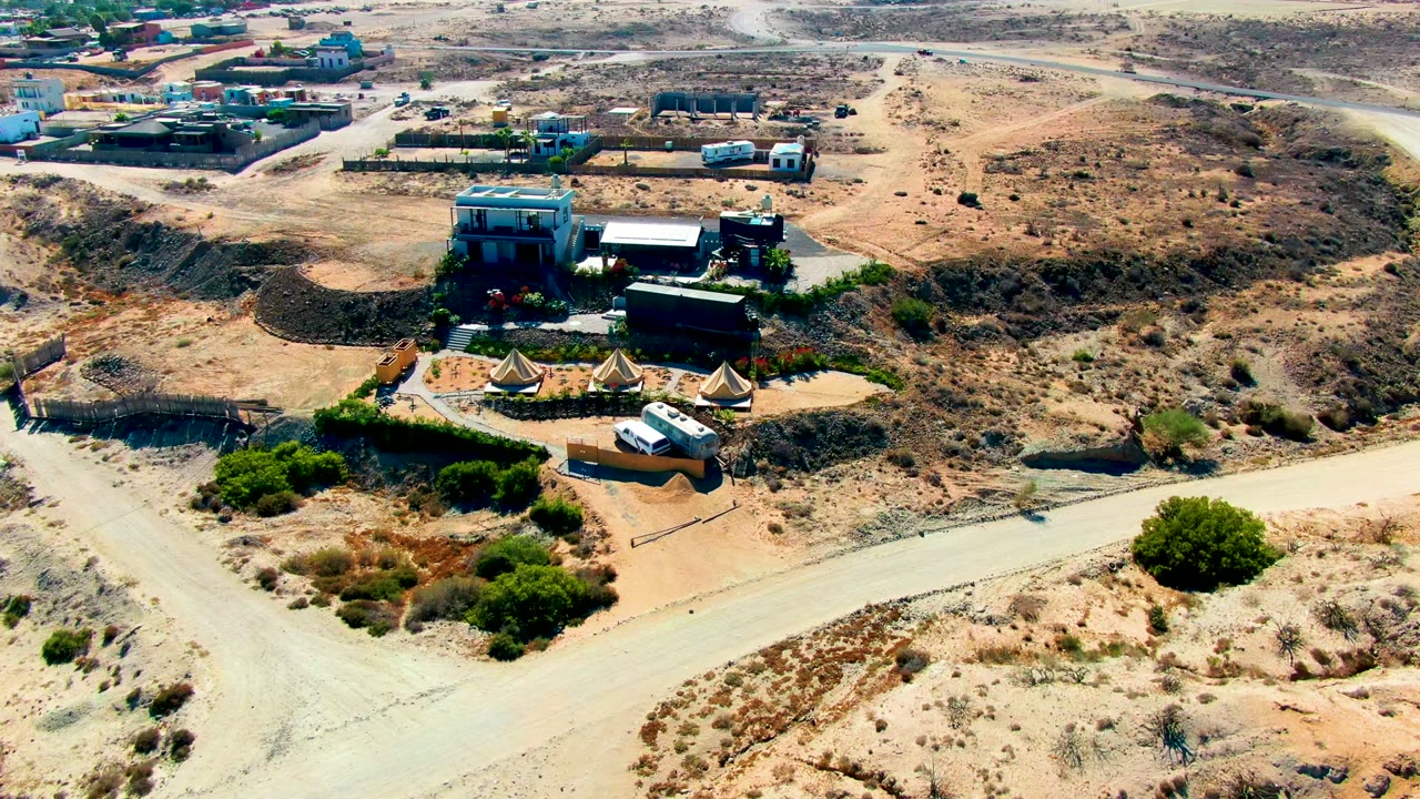 Drone view closer to tents, buildings, and a white van in desert terrain.