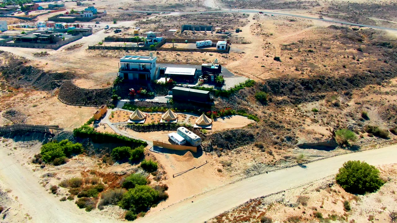 San Juanico, Baja California Sur aerial frame showing a cluster of tan tents below low buildings, pale access roads, scrub-covered desert, and no visible people.