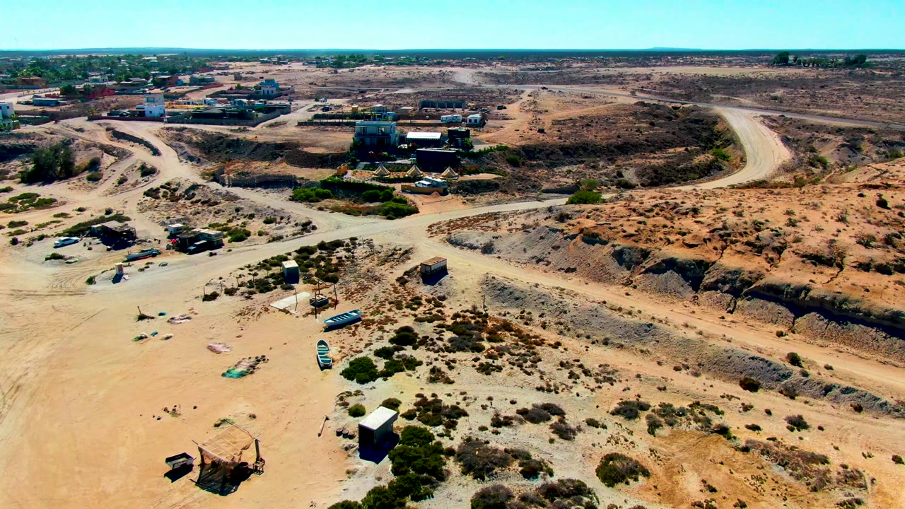 Aerial frame over San Juanico, Baja California Sur, showing sandy scrubland, pale dirt roads, small structures, parked vehicles, and no visible people.