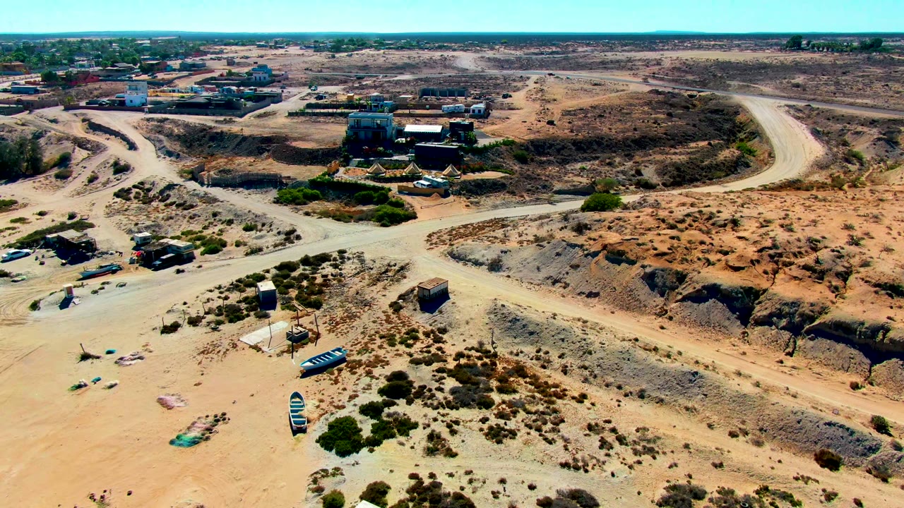 Drone frame over San Juanico showing a pale road bending through rocky orange-brown ground, with low structures, a few vehicles, sparse bushes, and no visible people.