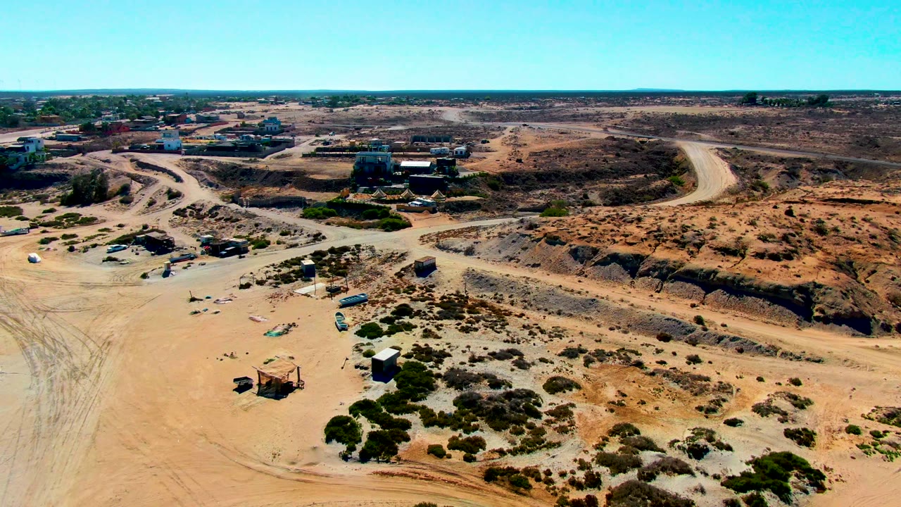 Second aerial frame in San Juanico, Baja California Sur with tire-marked sand in the foreground, shed-like structures below, winding dirt roads, distant buildings, and no visible people.