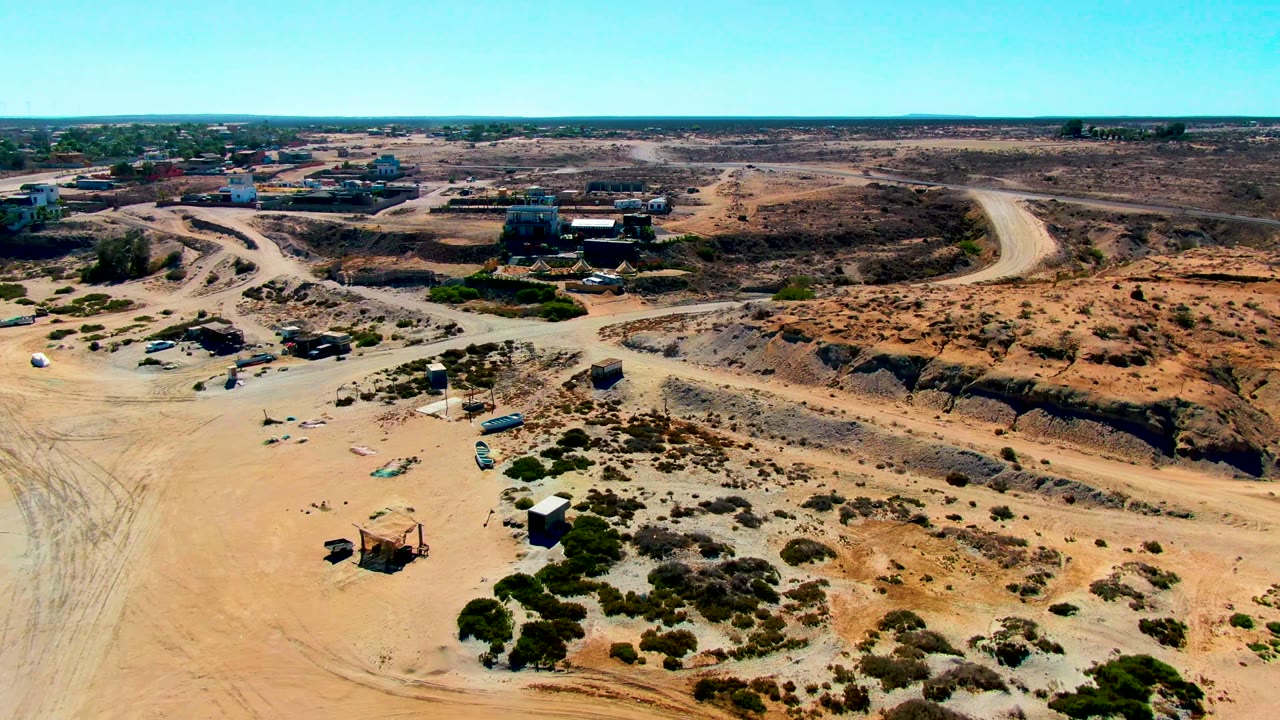 Mid-clip aerial frame over San Juanico, Baja California Sur showing a pale dirt road curving through an arid site, scattered equipment, low shrubs, distant buildings, and no visible people.