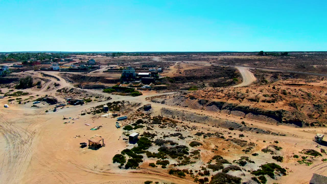 Aerial view over San Juanico, Baja California Sur, showing dirt roads, excavated tan earth, scrub vegetation, distant buildings, and no visible people.
