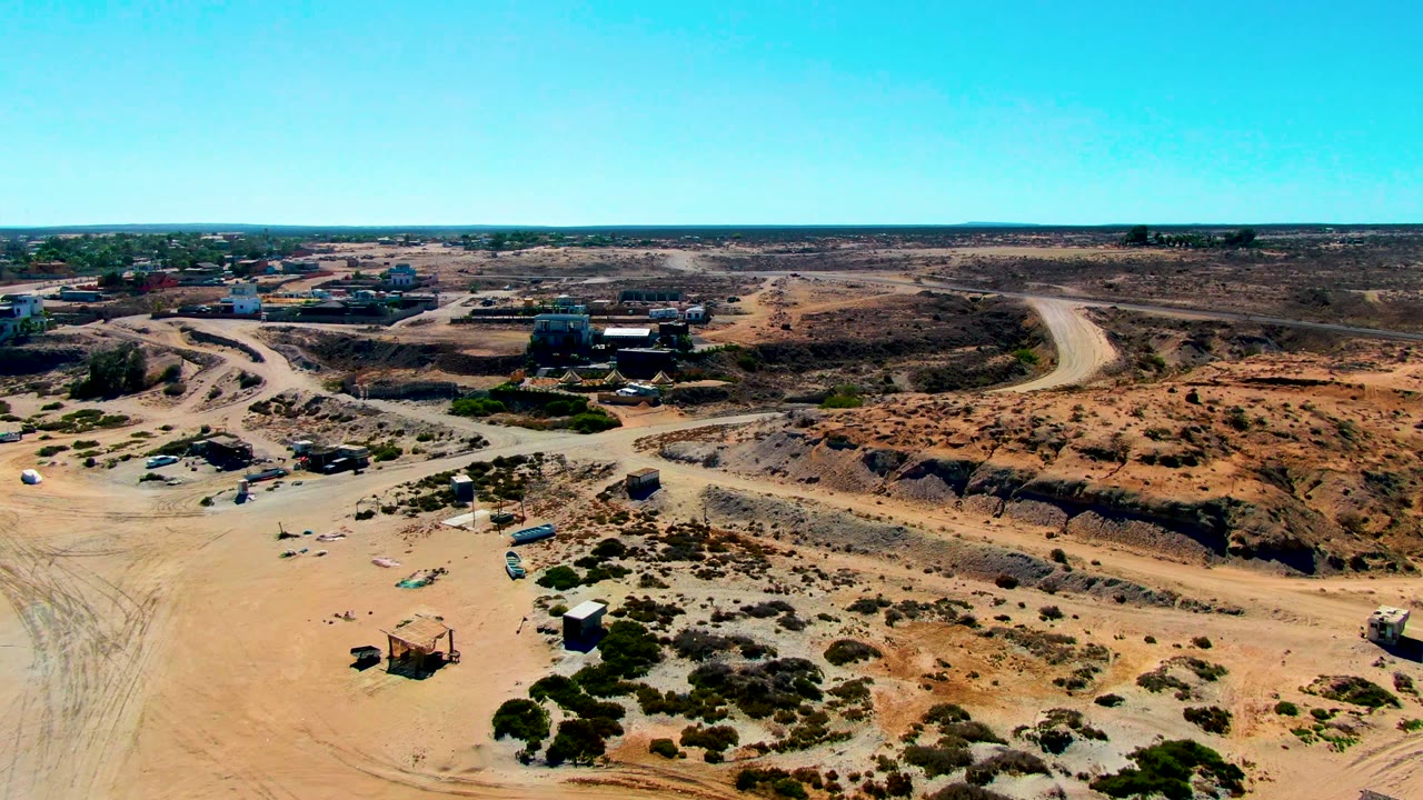Wide aerial frame over San Juanico, Baja California Sur, with dirt tracks crossing pale ground, brown excavation cuts to the right, buildings along the horizon, and no visible people.