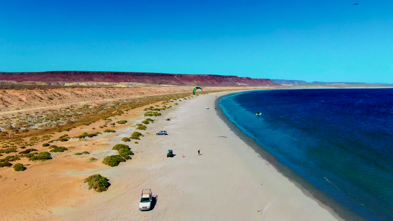 Aerial frame over San Juanico, Baja California Sur showing a wide sandy beach, blue ocean, desert bluffs, parked 4x4 vehicles, a green kite, and tiny people on shore.