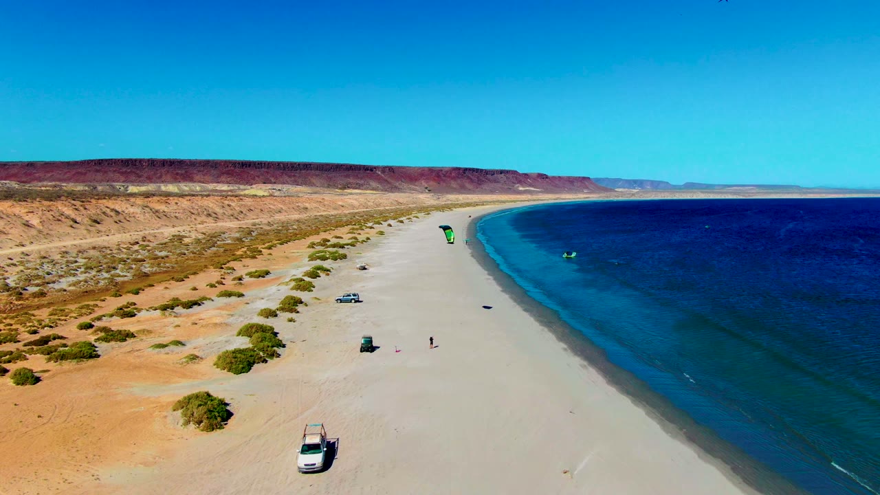 San Juanico aerial frame with the green kite closer to the shoreline, a small kite surfer on the water, parked vehicles on the sand, and red-brown desert bluffs in the background.