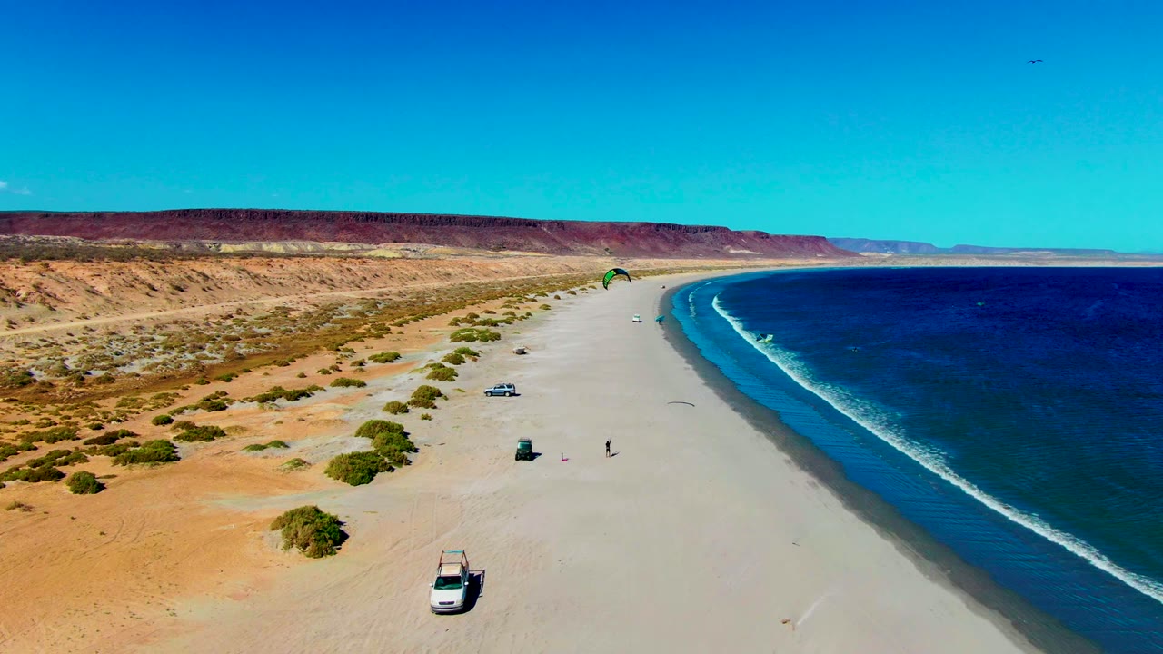 Wide aerial view over San Juanico beach in Baja California Sur with a pale sandy shoreline, deep blue ocean, desert scrub, a green kitesurfing kite, a white vehicle, and small people on the sand.