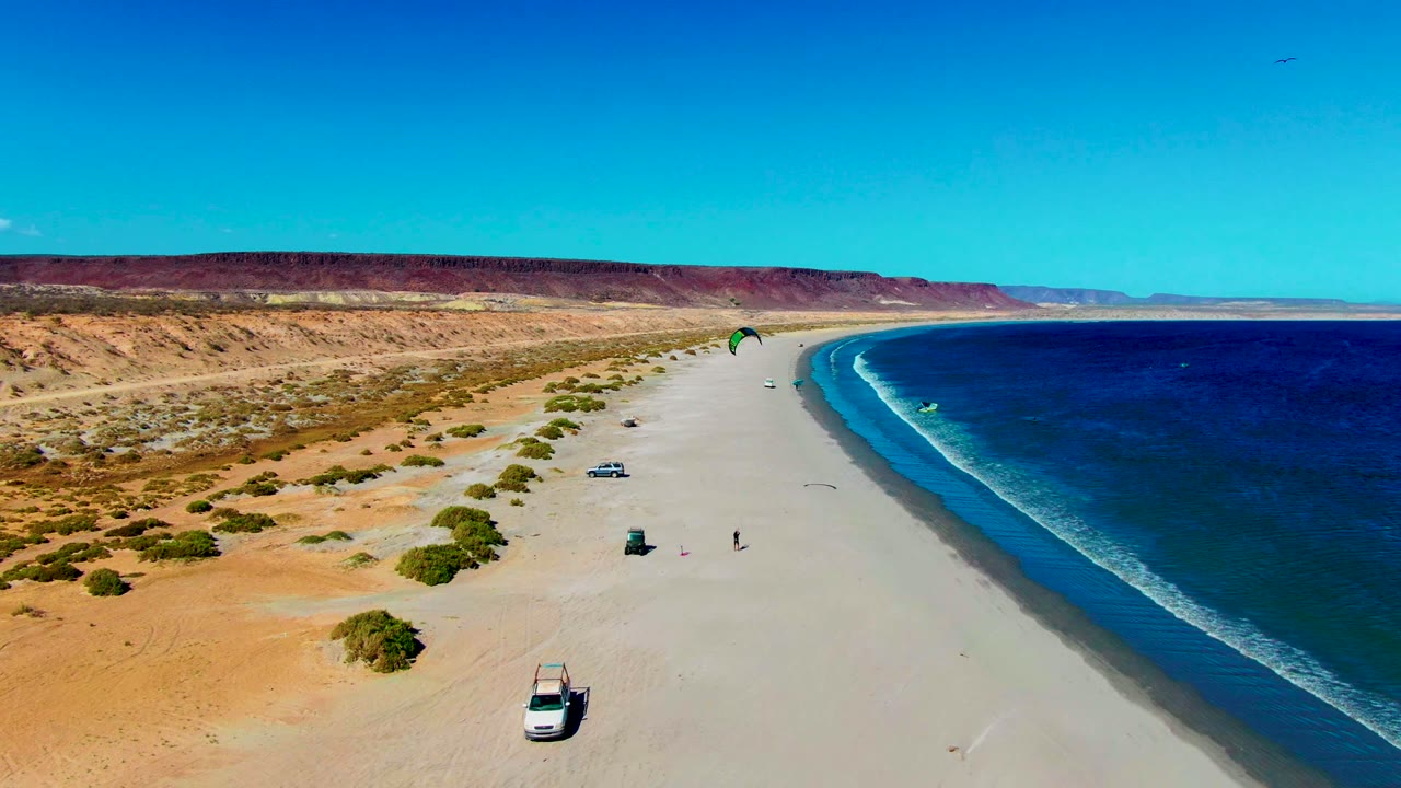 Aerial coastline with desert, beach vehicles, small people, and kitesurfing kite over the waterline.