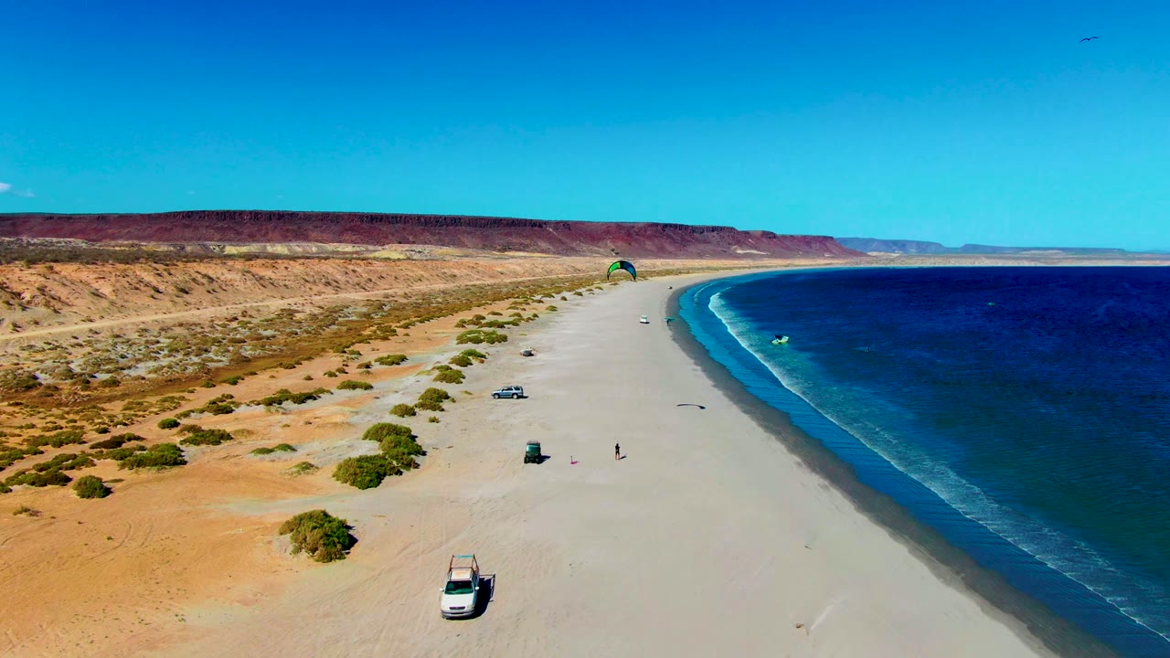 Aerial view of San Juanico's sandy coast with the green kitesurfing kite farther along the beach, a turquoise patch around a rider on the water, a white vehicle on the sand, and a red-brown desert ridge beyond.