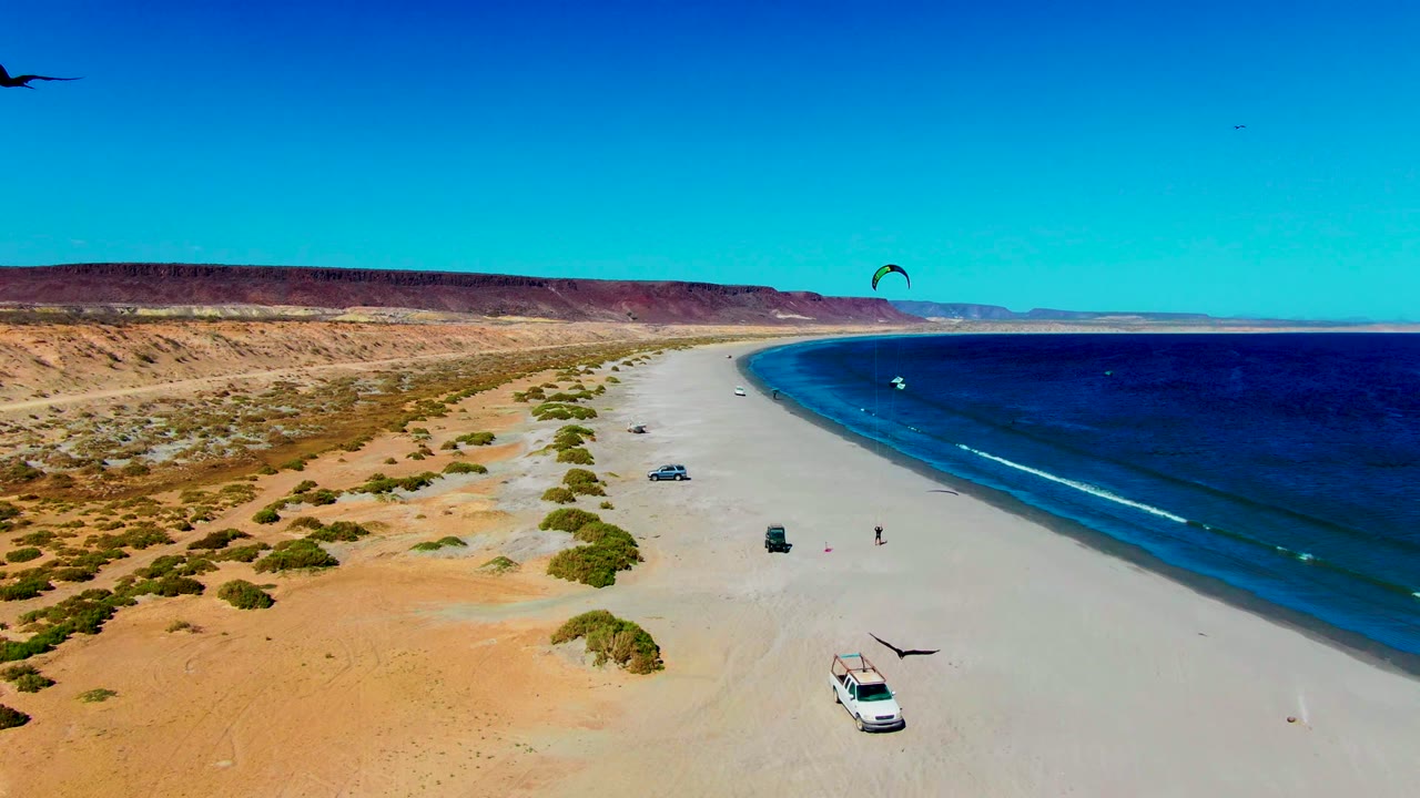 Aerial frame over San Juanico, Baja California Sur showing a curved sandy beach, desert scrub, blue ocean, a white vehicle near the bottom edge, smaller vehicles upshore, kite surfers offshore, and tiny people on the sand.