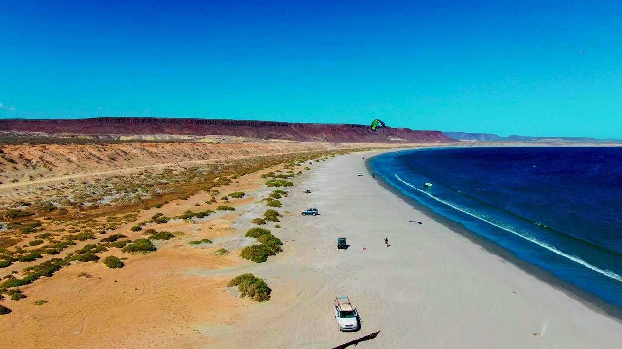 A wide San Juanico beach frame shows desert sand and scrub on the left, dark blue ocean on the right, vehicles spaced along the shore, a kite canopy above the water, and small people visible near the mid-beach area.