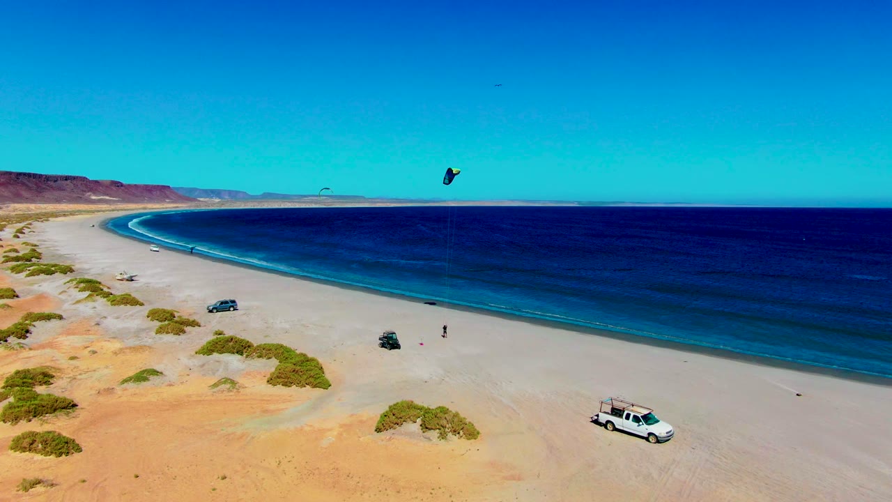 Aerial frame over San Juanico, Baja California Sur showing a sandy beach curving beside blue ocean, distant kite surfers, parked vehicles, desert scrub, and small visible people on the shore.