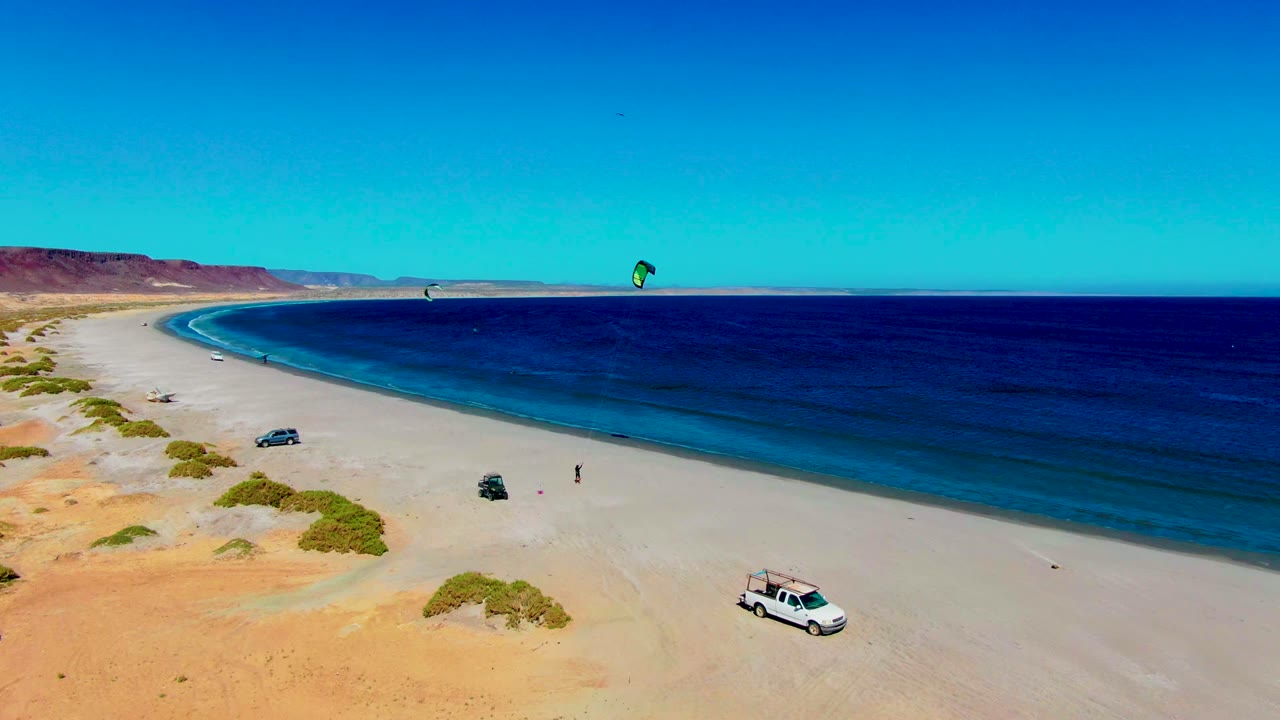 Aerial frame at San Juanico with a white van lower right, another vehicle near the beach center, kite surfers offshore, green scrub patches, distant cliffs, and small visible people on the sand.