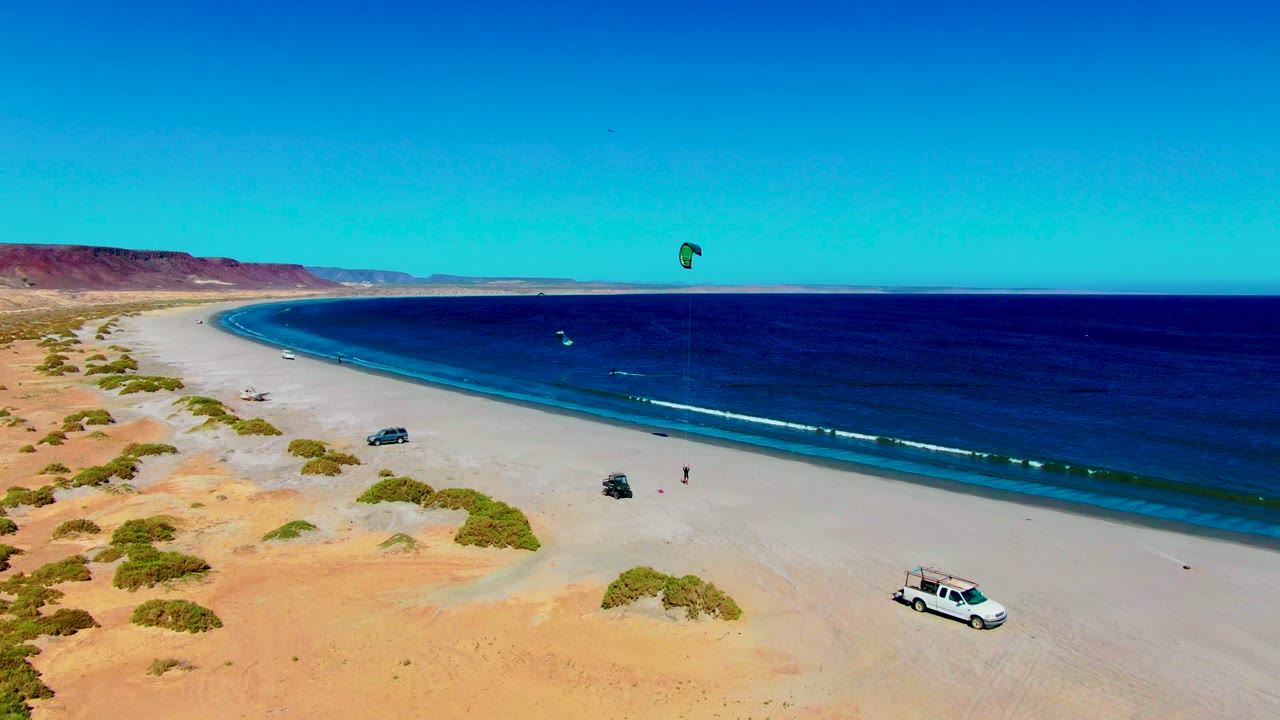 Aerial frame from San Juanico, Baja California Sur showing a broad sandy beach, blue ocean, one kite sail high in the sky, parked vehicles on the shore, and no visible people.