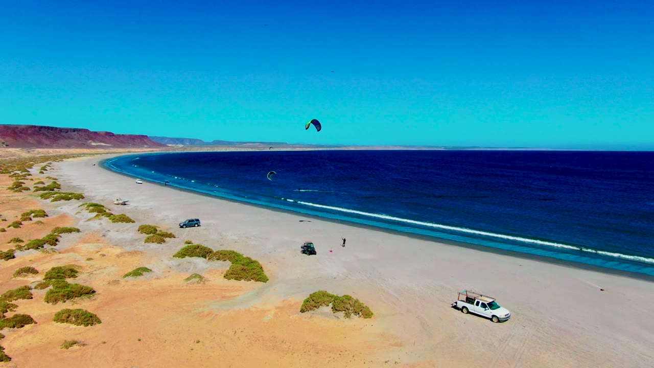 Wide beach aerial with two kite sails over the ocean and parked vehicles on sand.