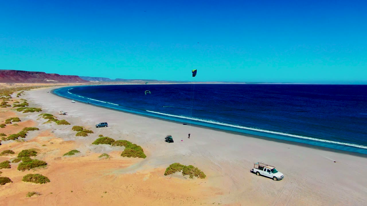 Aerial frame from San Juanico, Baja California Sur showing the white van farther right on the sand, a dark car near the beach center, distant kite sails above the ocean, and no visible people.