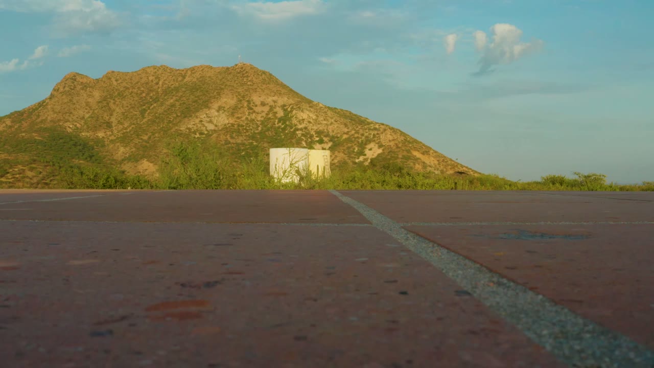 Opening frame of a low paved road view in Cabo Pulmo, Baja California Sur, with a pale line crossing the foreground toward green bushes, a white structure, and a rocky hill, no visible people.
