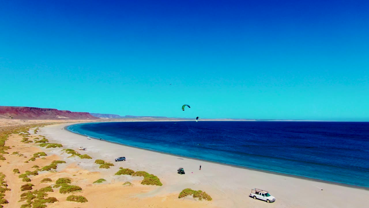 Aerial coastline with kite surfer offshore, parked vehicles on sand, dunes, and clear sky.