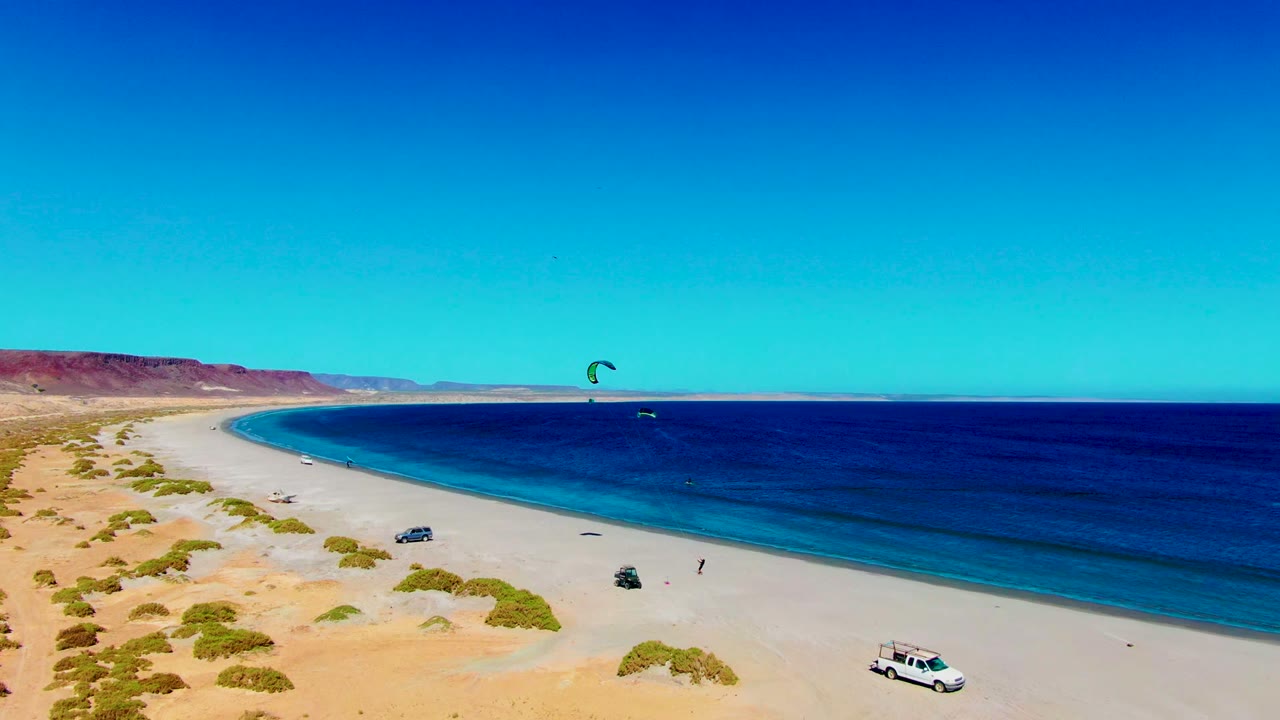 Wide aerial view of the San Juanico shoreline with the kite high over the ocean, vehicles parked on the open sand, desert dunes in the foreground, and no visible people.