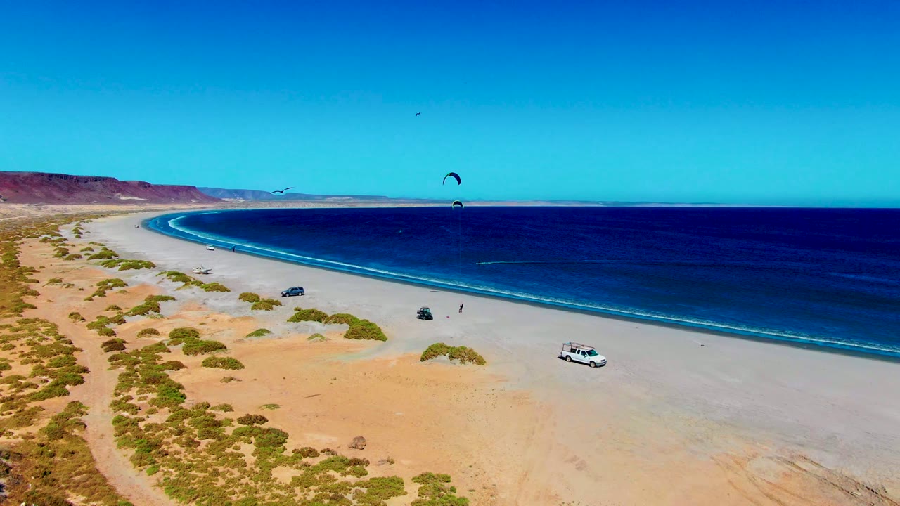 Aerial frame over San Juanico, Baja California Sur showing the curving sandy beach, deep blue ocean, desert shrubs, parked vehicles, and two kite canopies offshore.