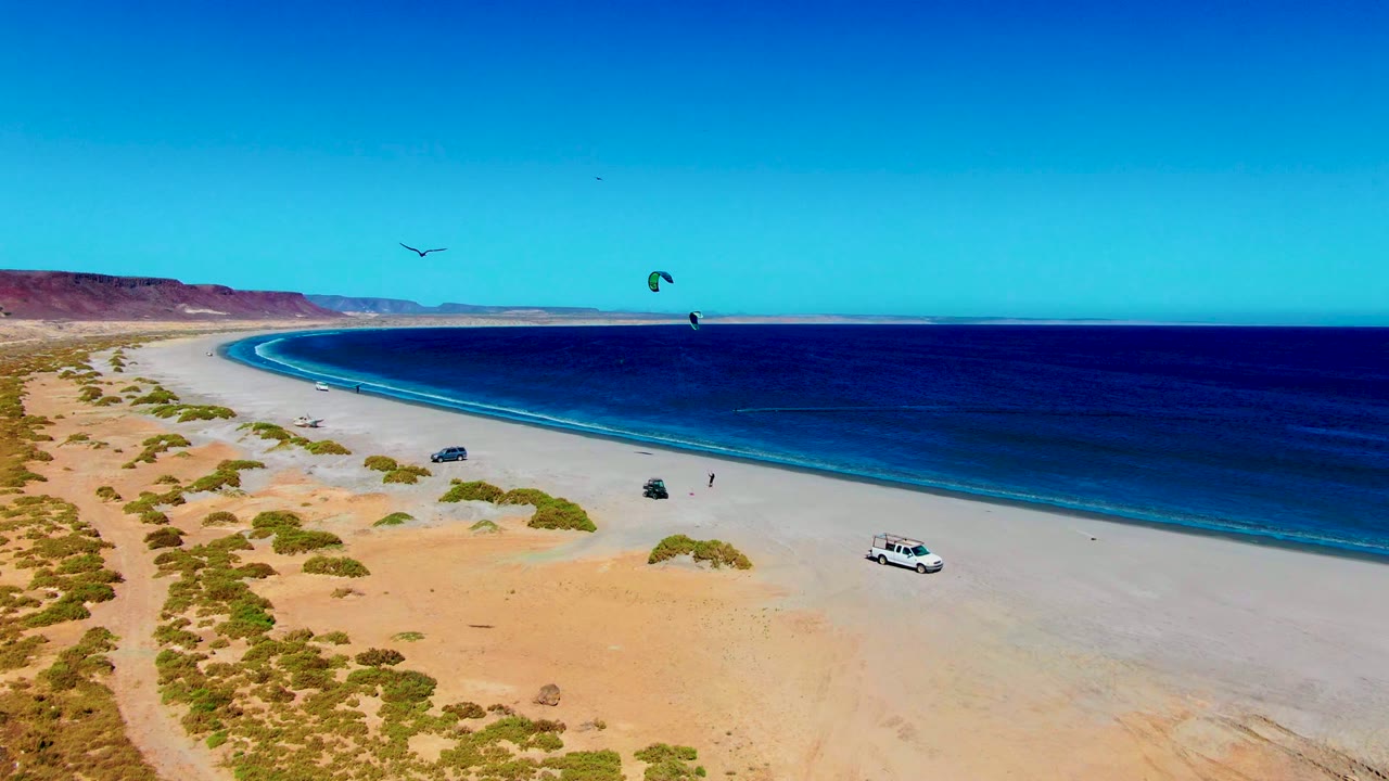 Wide aerial beach view with kite canopies over the water and vehicles on the sand.