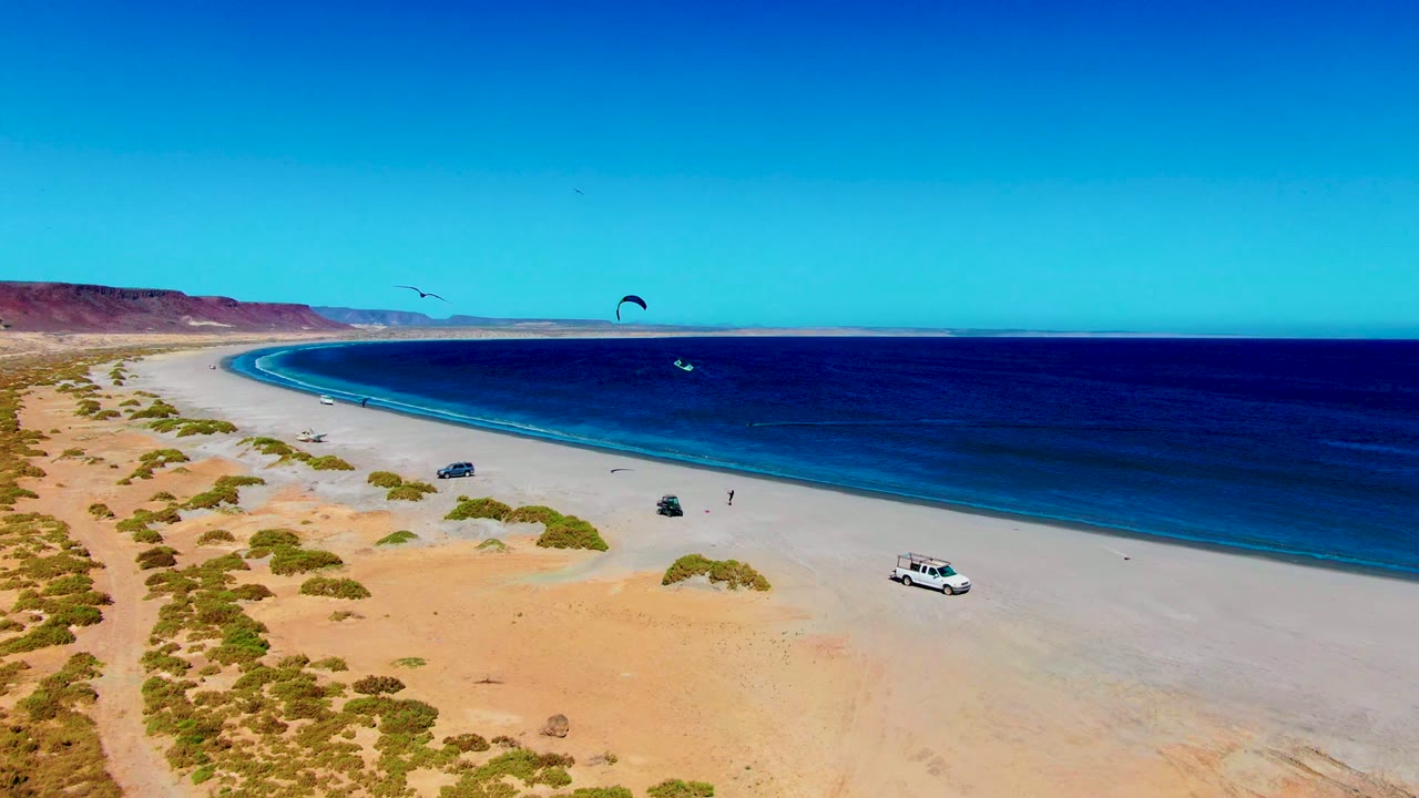 A wide San Juanico coastline frame showing pale sand, dark blue ocean bands, small kite canopies over the water, parked beach vehicles, and desert bluffs in the distance.