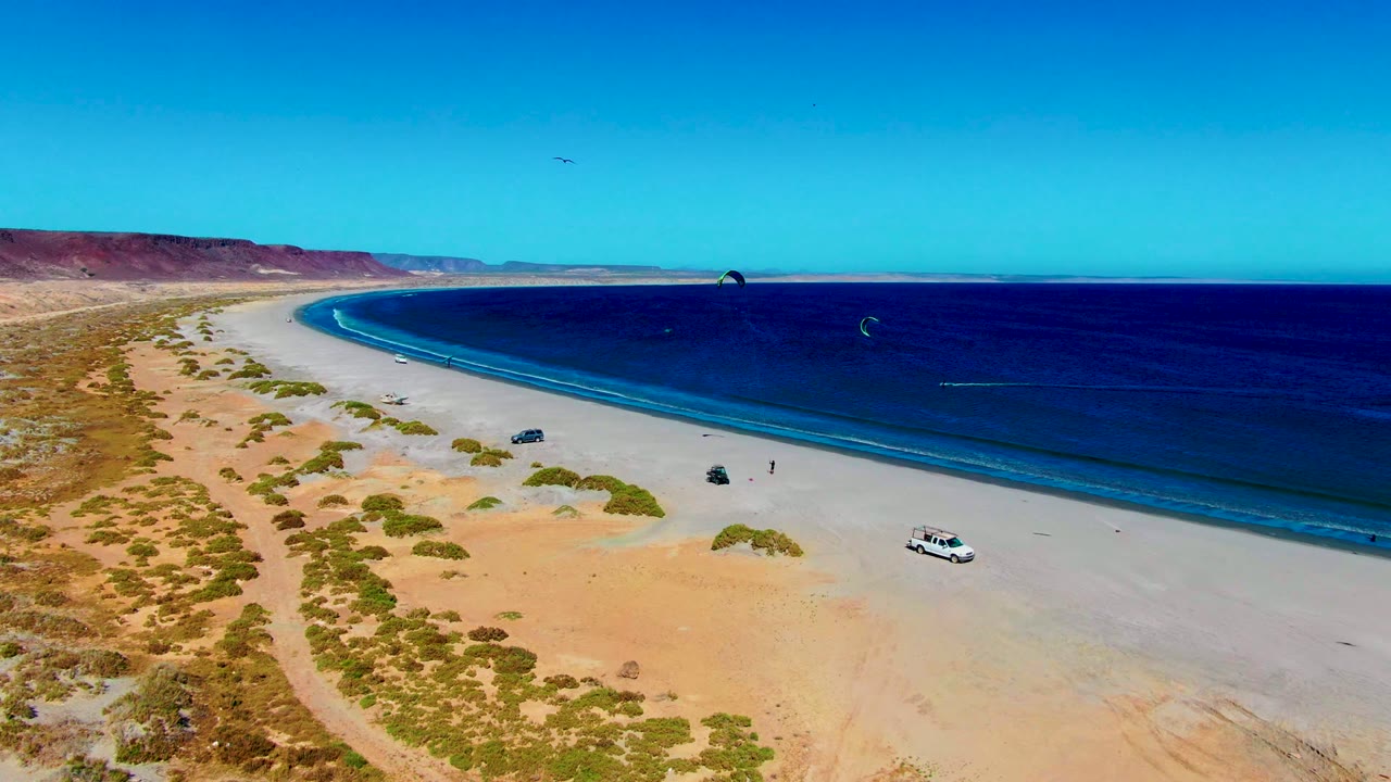 Drone view of curved beach with parked vehicles and kite sails over the water.