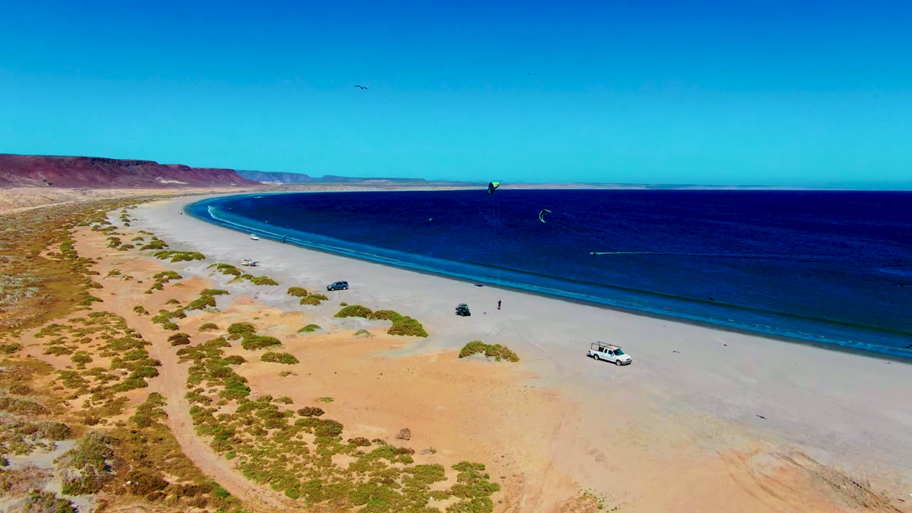 The San Juanico coastline fills the frame with tan sand, scattered shrubs, parked vehicles, two small people near the waterline, and kite surfers moving across the blue ocean under clear sky.