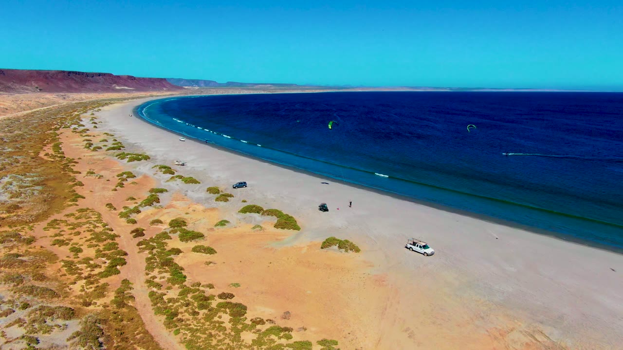 Aerial frame over San Juanico, Baja California Sur showing pale beach, desert scrub, parked vehicles, blue ocean, small kites offshore, and no visible people.