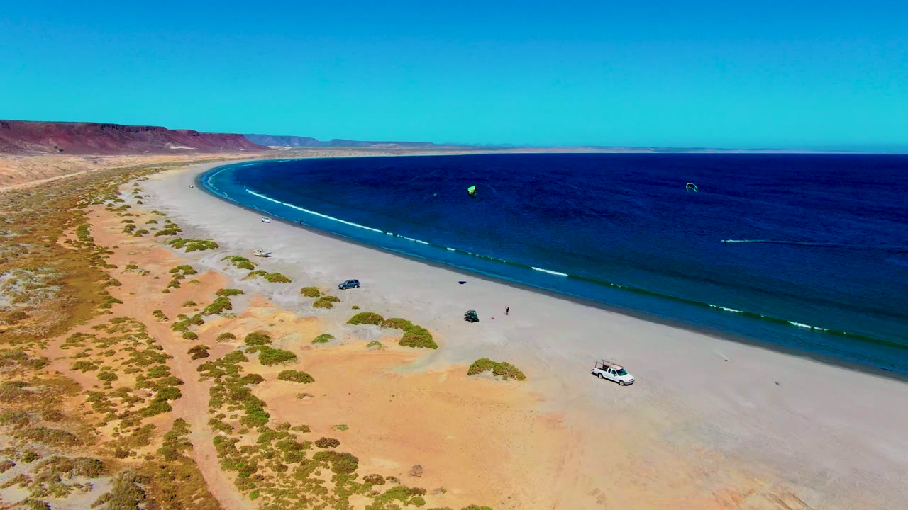 Wide drone view of curving beach with kites on the water and vehicles on sand.