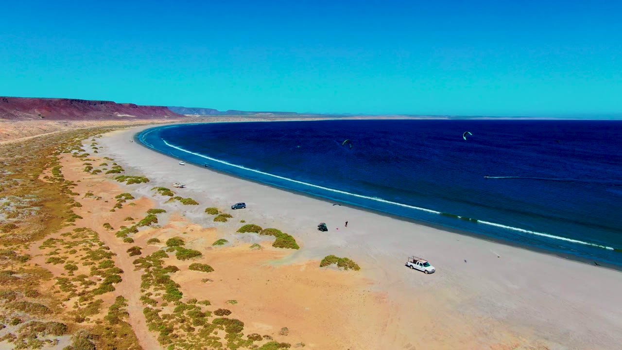 Drone-style frame over San Juanico showing a white vehicle near the lower beach, darker vehicles closer to the shore, kites on the ocean, and no visible people.