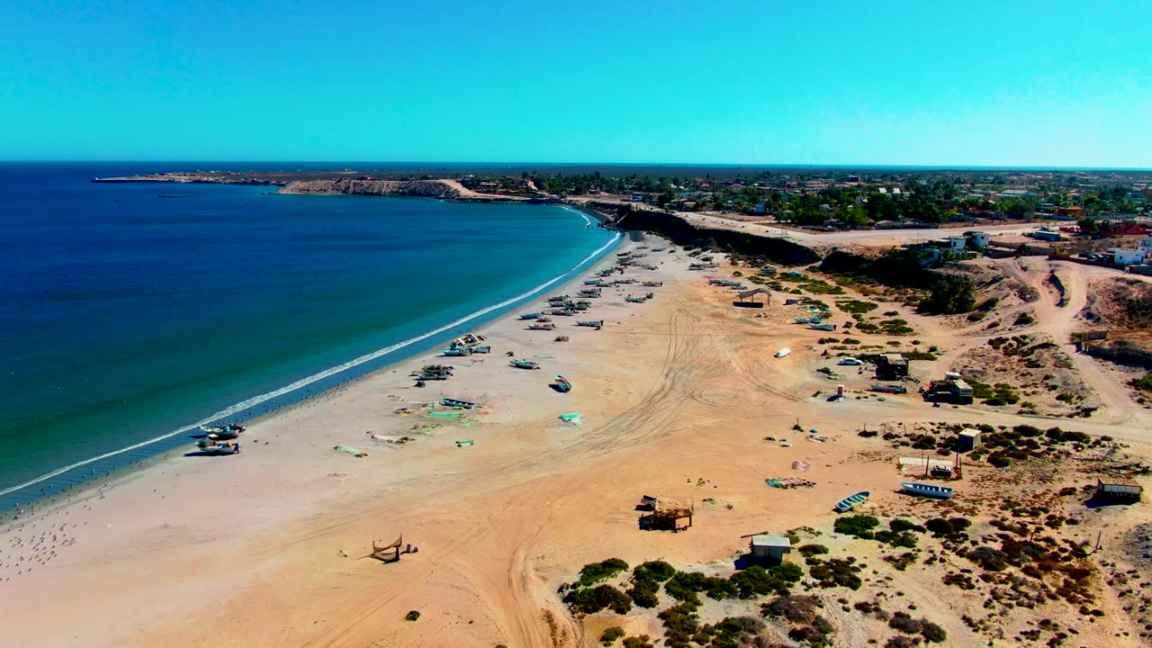 Aerial view over San Juanico, Baja California Sur, with calm blue ocean on the left, sandy beach and small boats along the shore, desert scrub inland, and no visible people.