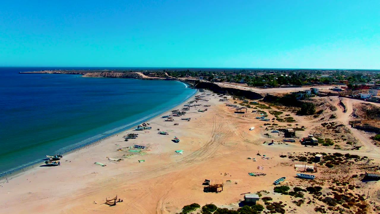 High aerial view of San Juanico beach with turquoise shallows, small boats spaced along the sand, darker desert vegetation to the right, distant structures, and no visible people.