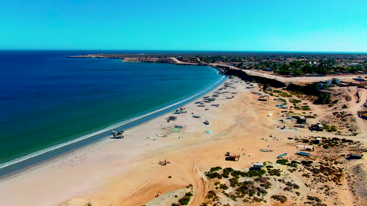 Aerial frame over San Juanico, Baja California Sur, showing blue ocean on the left, a broad sandy beach with small boats, low buildings beyond the shore, and no visible people.