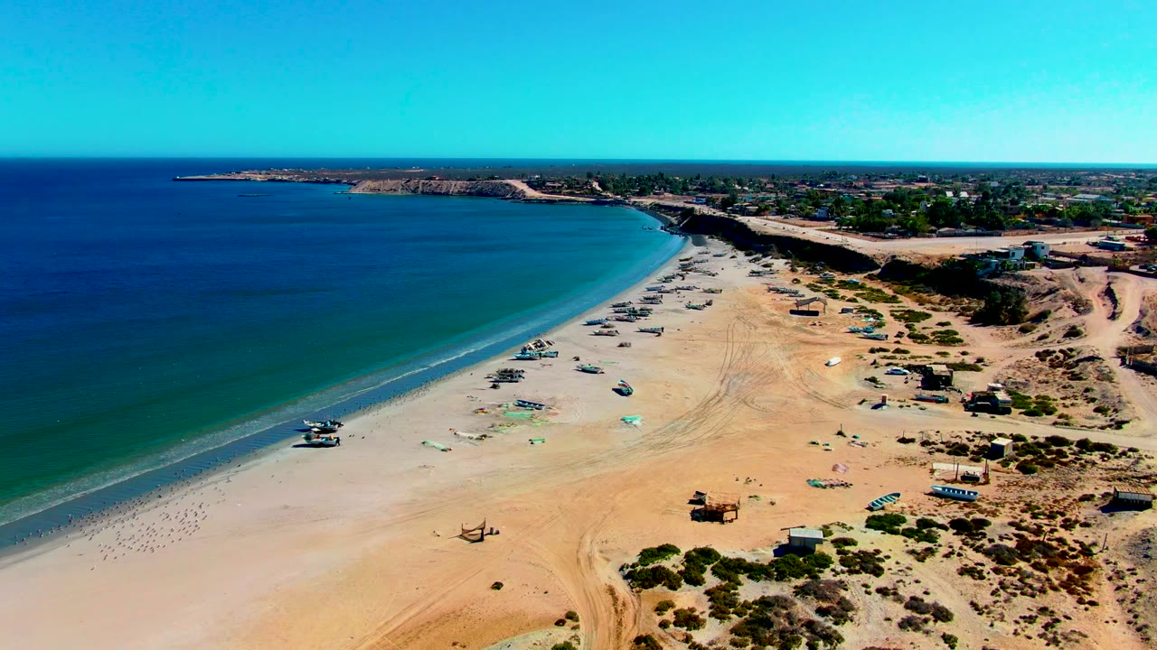 The San Juanico coastline remains in a high aerial composition, with calm surf tracing the beach, clustered small boats on the sand, inland buildings, sparse shrubs, and no visible people.