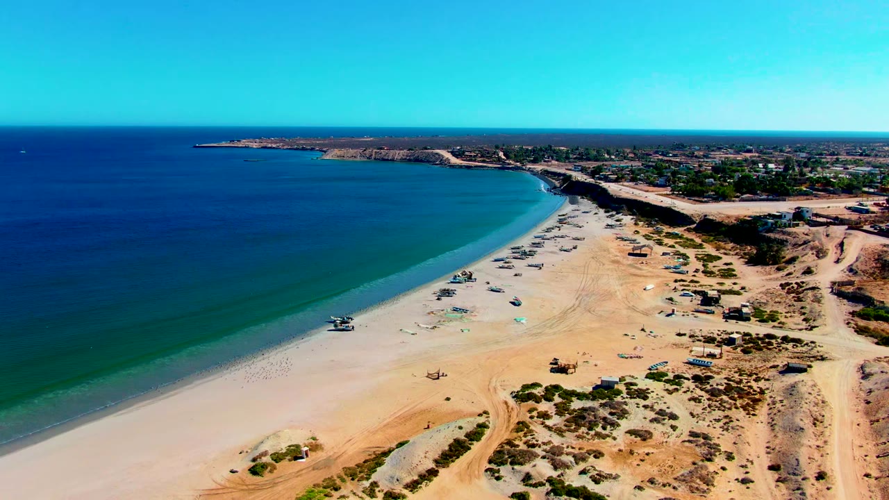 Aerial frame over San Juanico, Baja California Sur, with deep blue ocean on the left, pale sand curving along the shore, small boats near the waterline, scrubby dunes to the right, and no visible people.