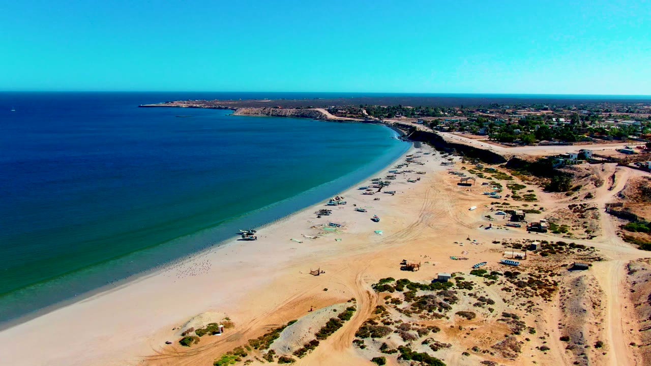 High coastal view with turquoise shallows, sand, boats, dunes, and distant buildings.