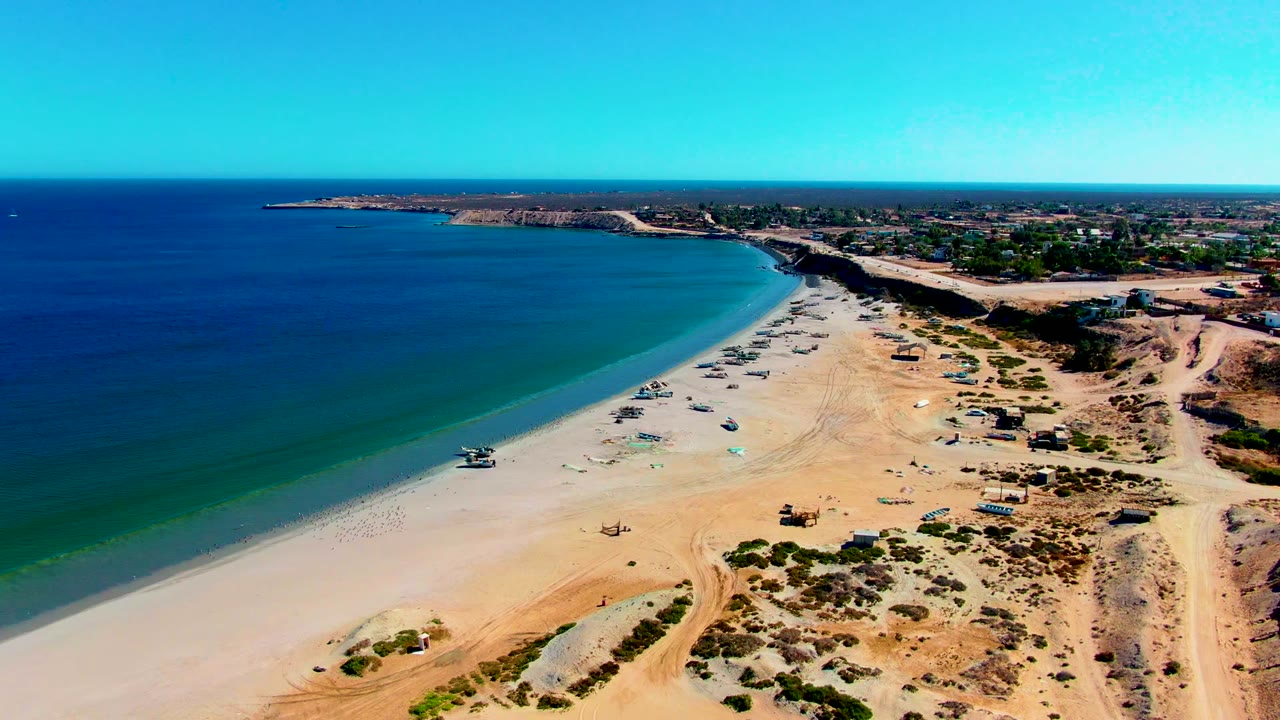 A wide aerial frame in San Juanico shows the shoreline running diagonally through the image, anchored boats on open sand, dry dunes and sparse shrubs in the foreground, ocean to the left, and no visible people.