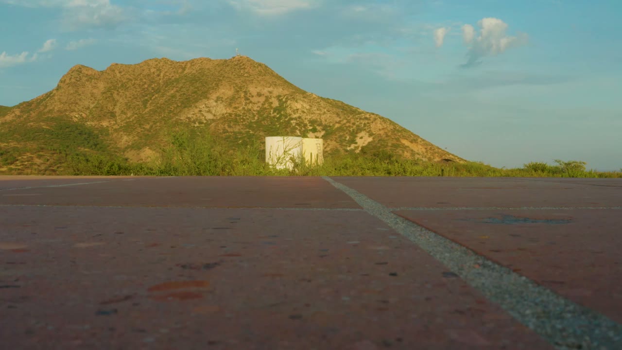 At 0:00, a low roadside view in Cabo Pulmo, Baja California Sur shows a pale line on reddish pavement leading toward brush, a small pale structure, a mountain, blue sky, and no visible people.