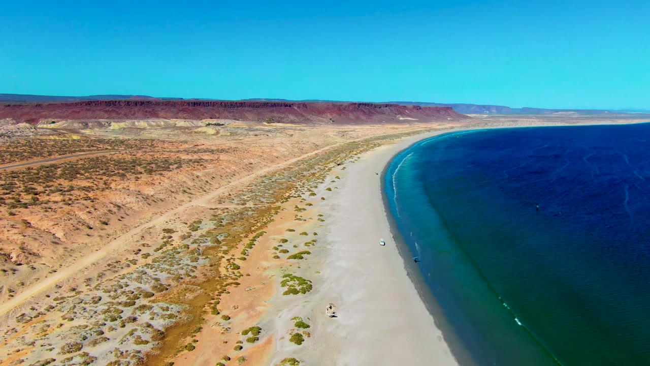 Aerial frame over San Juanico, Baja California Sur showing pale sand, sparse desert scrub, deep blue ocean, red-brown hills, clear sky, and no visible people.